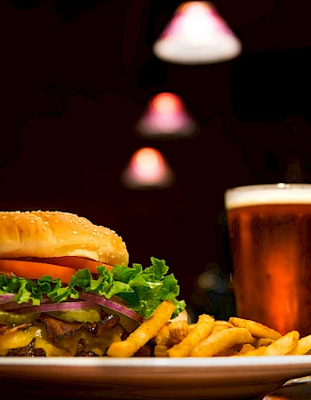 A close-up of a plate with a burger, fries, and a glass of beer under warm lighting in a dimly lit setting.