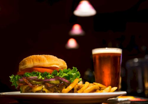 A cheeseburger with lettuce, tomato, and onions, served with fries, and a glass of beer on a table with dim, warm lighting.
