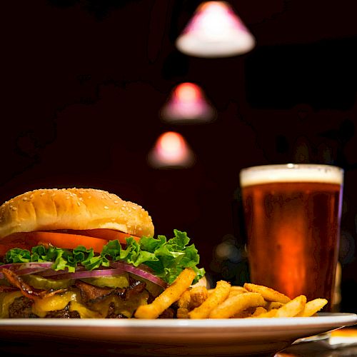 A cheeseburger with lettuce, tomato, and onions, served with fries, and a glass of beer on a table with dim, warm lighting.